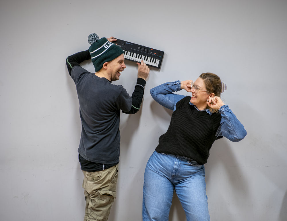 Två personer står mot en vägg, en håller ett litet keyboard och den andra håller för öronen och ler.
