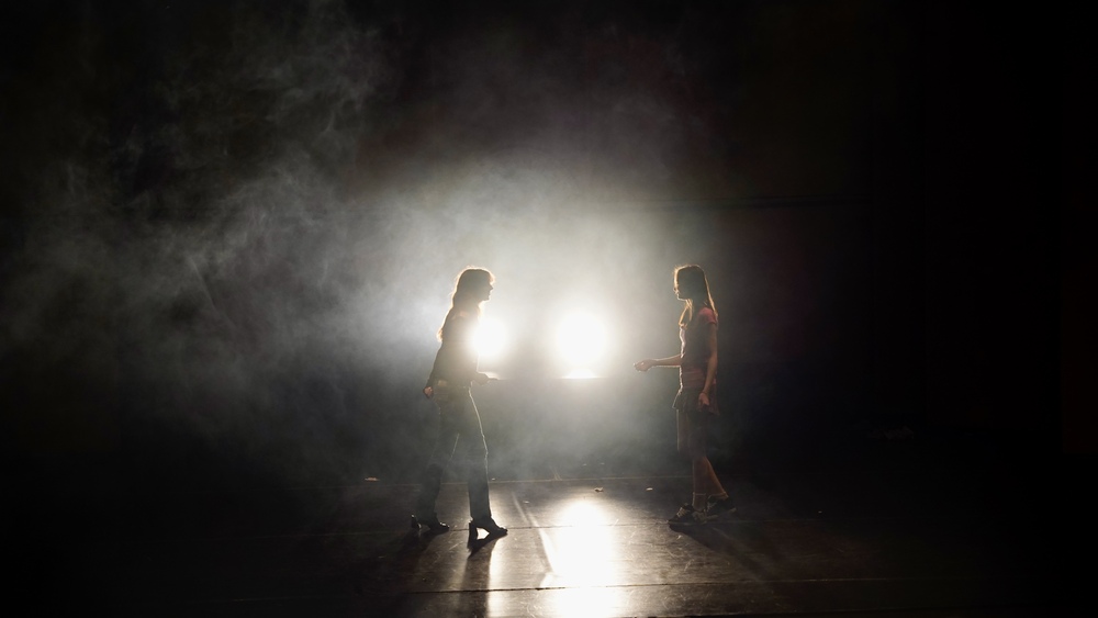 Två personer står mittemot varandra på en mörk scen med starkt motljus och rök.