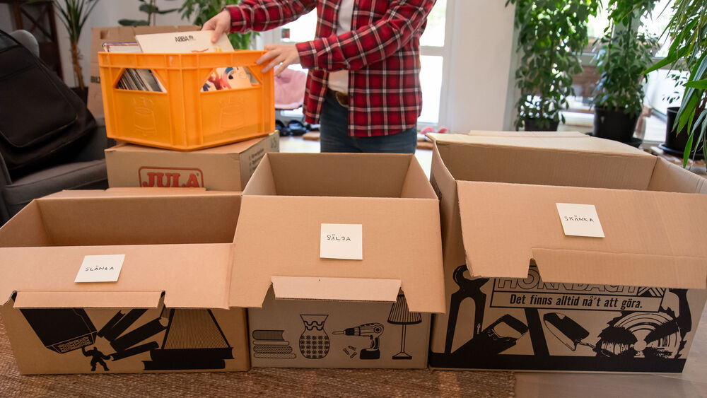 En person packar eller sorterar saker i flyttkartonger och plastlådor i ett ljust rum.