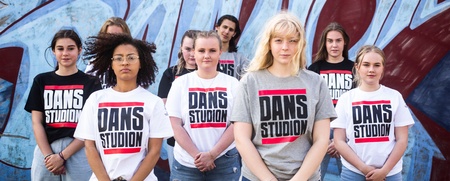 En grupp unga personer står framför en graffitivägg, alla bär t-shirts med texten 'DANS STUDION'.