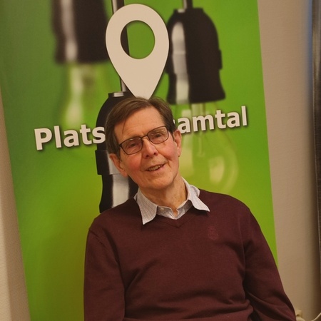 Man med glasögon sitter vid ett bord framför en grön skylt med texten 'Plats för samtal'.