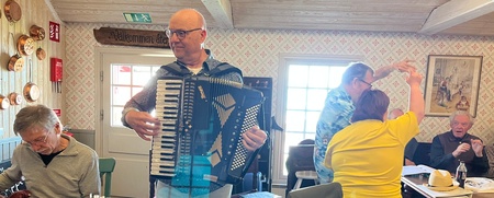 En man spelar dragspel i ett ljust rum, andra personer sitter, spelar gitarr eller dansar.