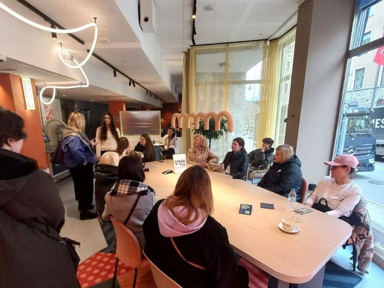 En grupp människor sitter och står runt ett bord i ett modernt café med stora fönster.
