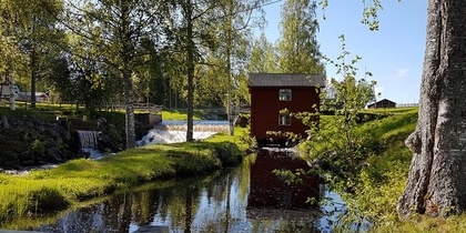 En röd stuga vid en liten fors och damm, omgiven av grönskande träd och gräs under en klarblå himmel.