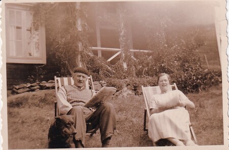 Ett äldre par sitter i solstolar utomhus med en hund, framför ett hus, i en gammaldags sepia-tonad bild.