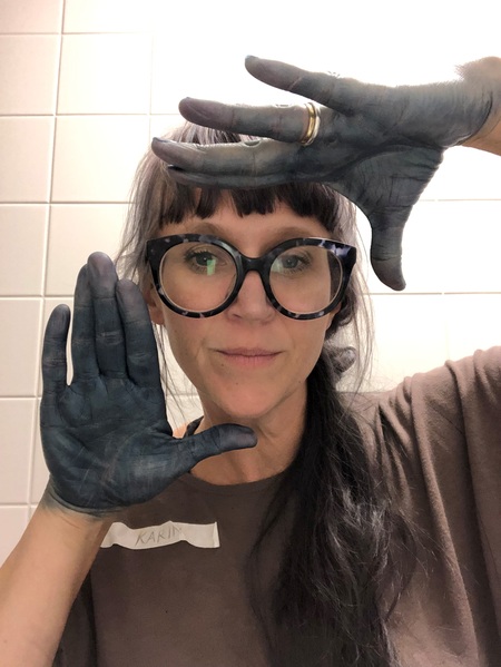 Kvinna med stora glasögon visar sina blåfärgade händer framför ansiktet, står mot vit kakelvägg.