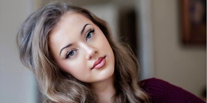 Ung kvinna med långt lockigt hår sitter avslappnat inomhus och tittar mot kameran.