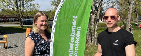 Två personer står vid ett informationsbord utomhus med en grön flagga från Studieförbundet Vuxenskolan.