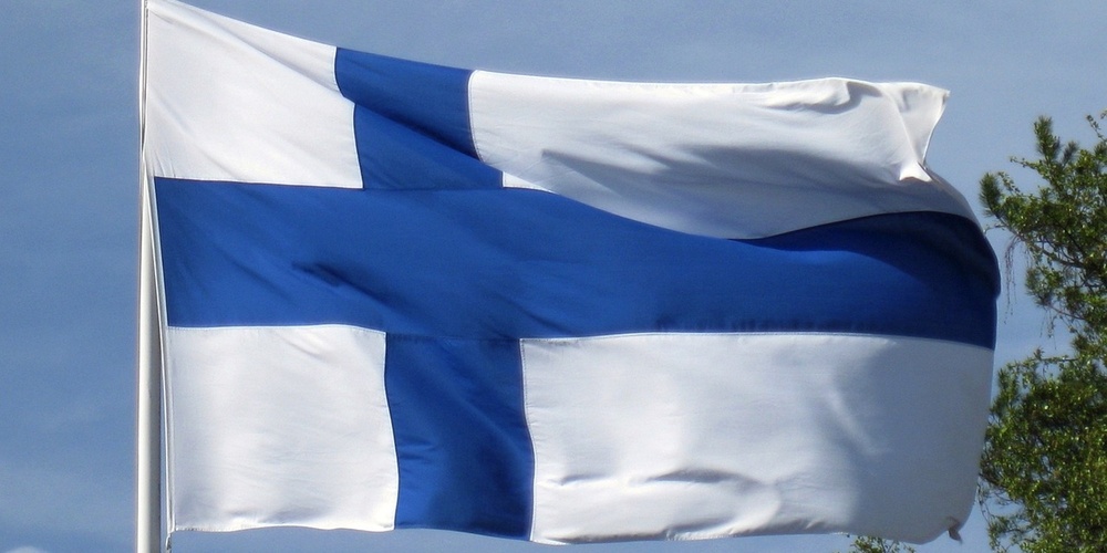 Finlands flagga vajar i vinden mot en blå himmel med några träd i bakgrunden.