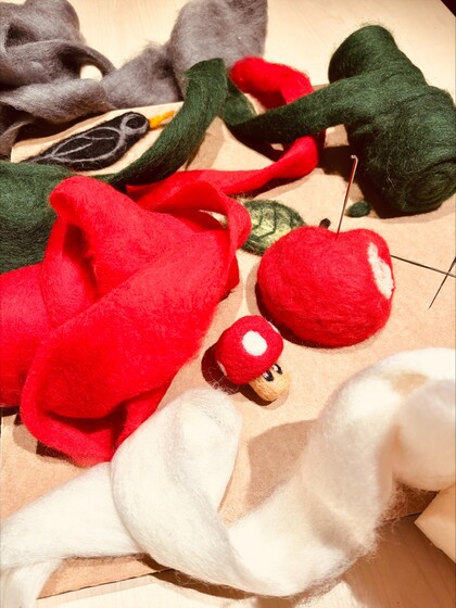 Färgglada ullbitar och tovade figurer, som ett äpple och en svamp, ligger på ett bord med tovningsnålar.