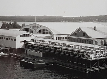 Svartvit bild av en industribyggnad vid vattnet med stor skorsten och bryggor.