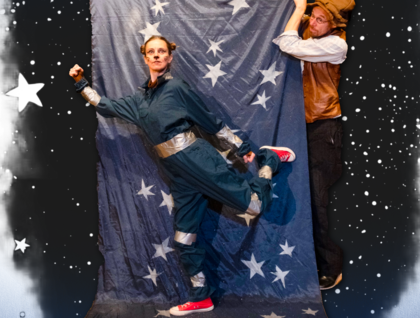 Två personer poserar framför ett blått tyg med vita stjärnor, en i overall och en i väst, mot en stjärnhimmelbakgrund.