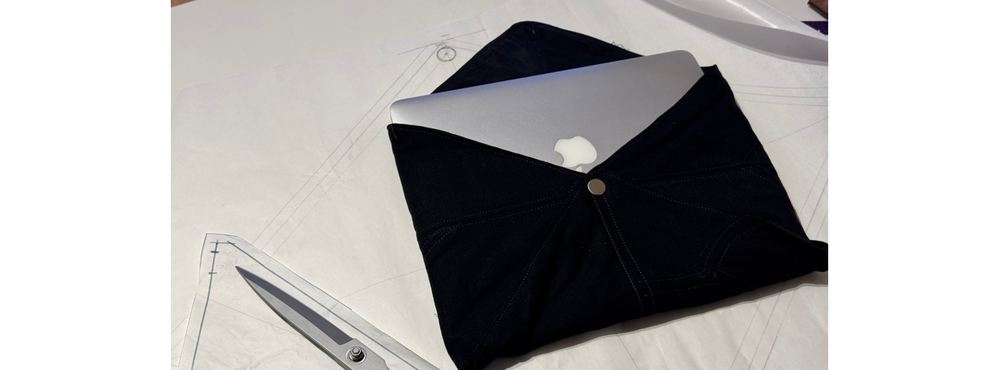 En laptop sticker upp ur ett svart tygfodral, bredvid en sax och mönsterpapper på ett bord.