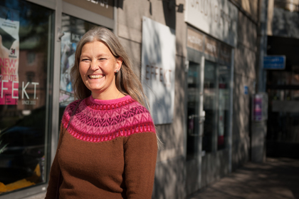 Leende kvinna med långt grått hår och brun tröja med rosa mönster står utomhus vid en butik.