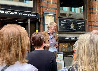 En man håller papper och pratar inför en grupp människor utanför en restaurang i stadsmiljö.