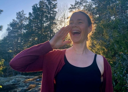 Person utomhus i skogsmiljö, håller ena handen vid ansiktet, ansiktet är suddat.