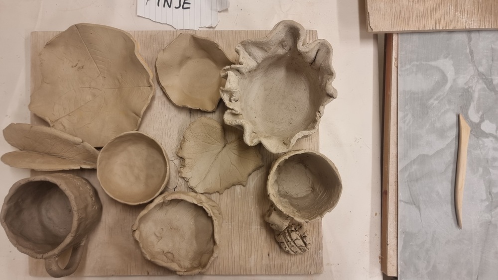 Olika handgjorda keramikföremål i lera, inklusive skålar, blad och en mugg, ligger på en träplatta.
