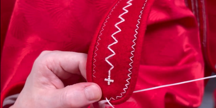 En hand broderar vita mönster på ett rött tyg med dekorativa sömmar och pärlor.