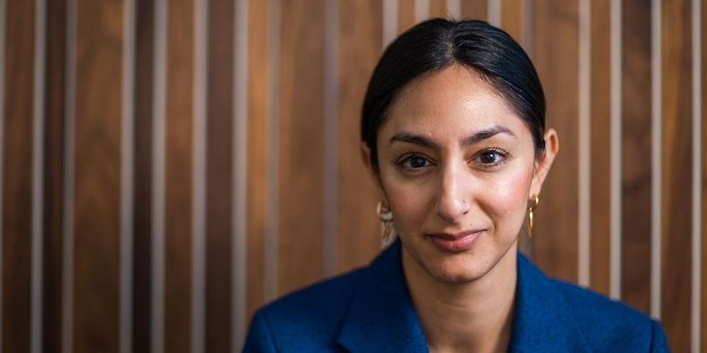 Kvinna med mörkt hår och blå kavaj sitter framför en trävägg med vertikala ränder.