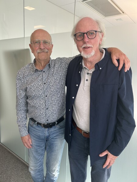 Två äldre män står bredvid varandra, den ena med armen runt den andra, i ett ljust kontorsrum.
