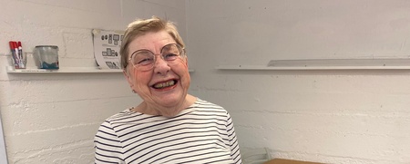 Leende äldre kvinna med randig tröja och taxmotiv står i ett ljust rum med bord och stolar.