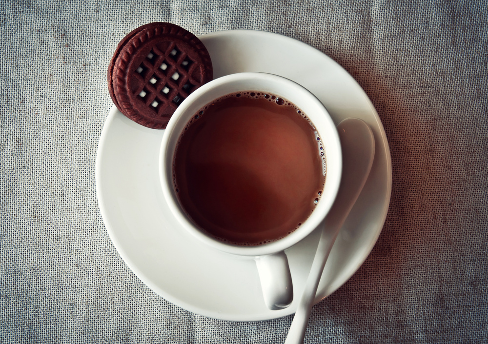 En kopp varm choklad på ett vitt fat med en chokladkaka och en vit sked, ovanifrån.