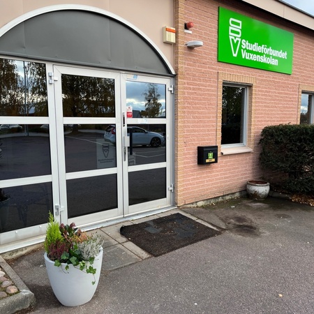 Ingång till en byggnad med glasdörrar och en grön skylt med texten 'Studieförbundet Vuxenskolan'.