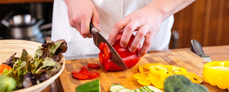 En person skär röda paprikor på en skärbräda omgiven av olika grönsaker i ett kök.