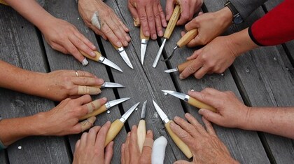 Flera händer med snidade knivar pekar mot mitten på ett träbord, några har plåster på fingrarna.
