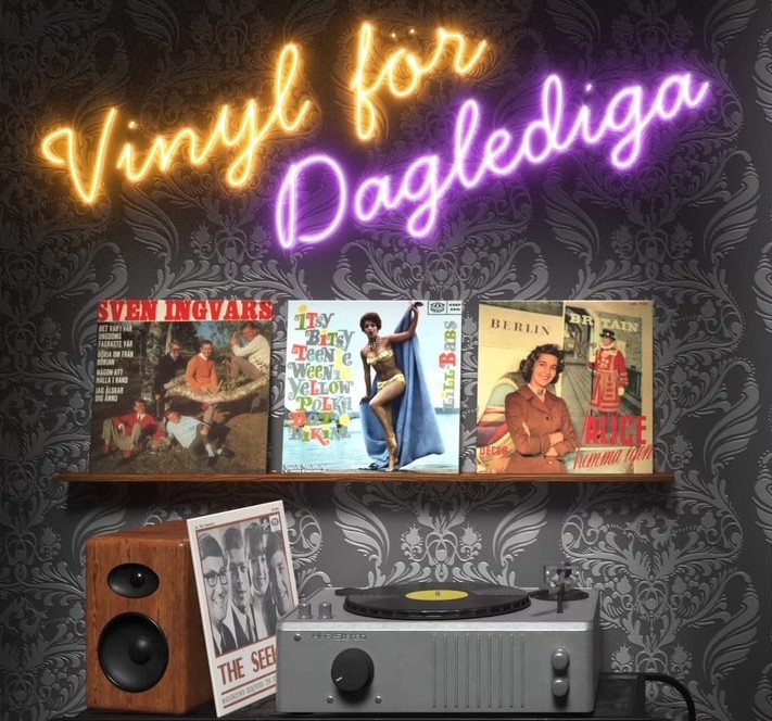 En hylla med vinylskivor, en skivspelare och högtalare under en neonskylt med texten 'Vinyl för Daglediga'.
