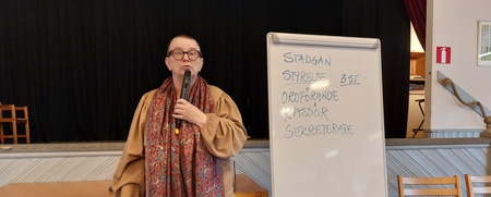 Person står vid ett blädderblock i en sal och håller en mikrofon. Text på blocket handlar om styrelseposter.