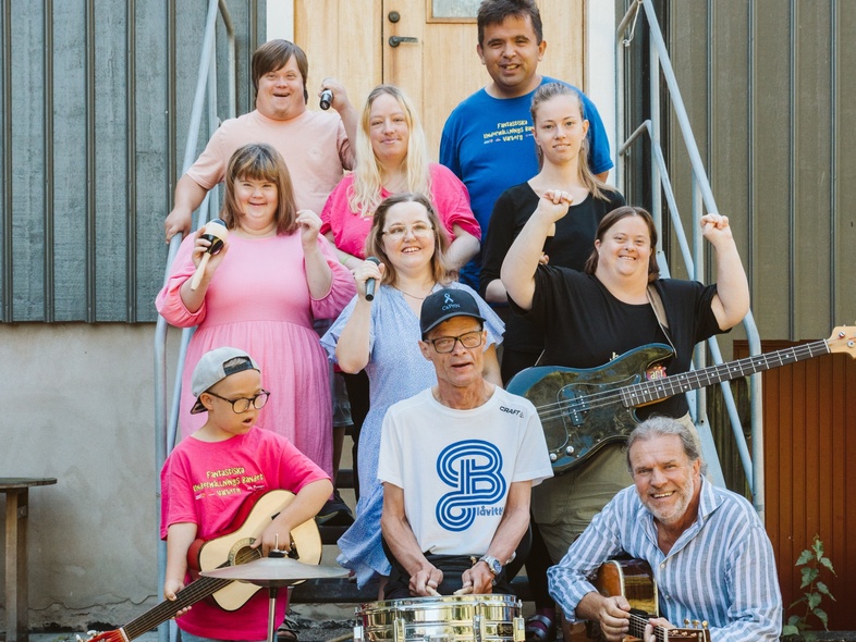 En grupp människor poserar utomhus framför ett hus, flera håller musikinstrument.