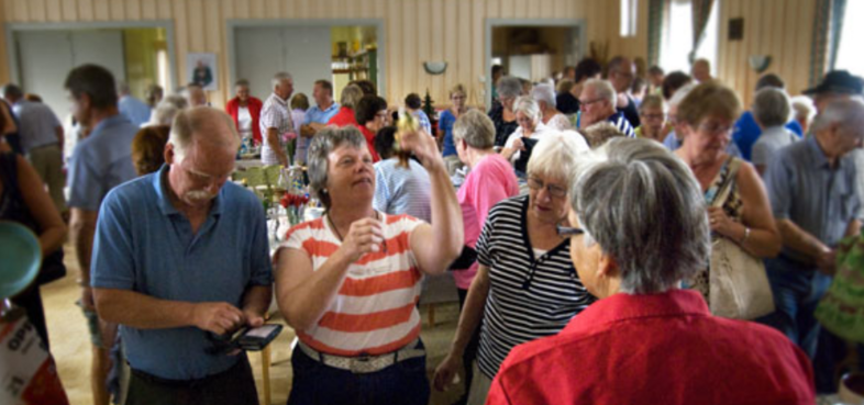 En grupp äldre personer samlade i en ljus lokal, samtalar och tittar på föremål vid ett bord.