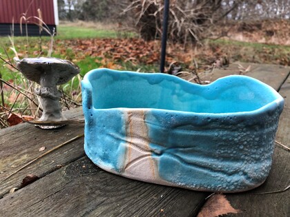 En blå keramikskål och en liten svampformad skulptur står på ett träbord utomhus med höstlöv runtom.