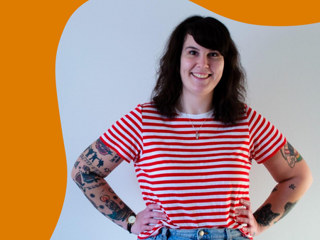Kvinna med mörkt hår, randig rödvit tröja och tatueringar står leende mot en vit och orange bakgrund.