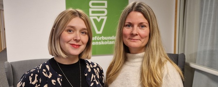 Två kvinnor står bredvid varandra inomhus framför en grön skylt med text.