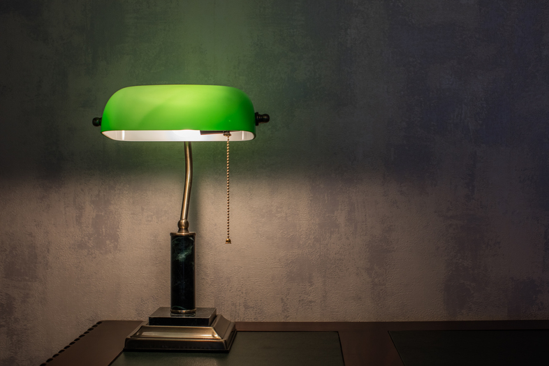 En klassisk skrivbordslampa med grön skärm står tänd på ett skrivbord mot en grå vägg.