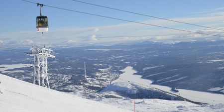 Landskapsvy över snöklädda fjäll med en linbana och utsikt över en frusen sjö och skog.
