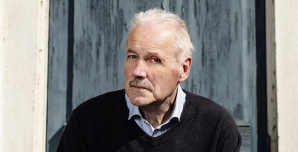 En äldre man med vitt hår sitter utomhus framför en blå dörr. Hans ansikte är suddat.