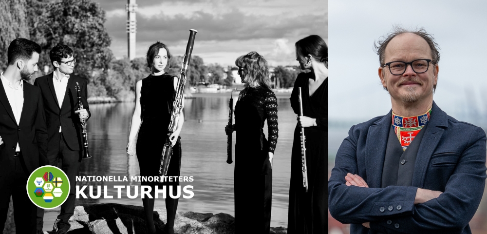 En grupp musiker med blåsinstrument står vid vattnet, och en man med glasögon och folkdräkt står bredvid.