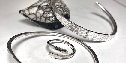 Tre silversmycken, två armband och en ring, ligger på ett vitt underlag med en svartvit sten i bakgrunden.
