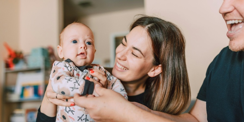 En mamma håller upp sin bäbis, två armar till från en andra förälder syns i förgrunden.