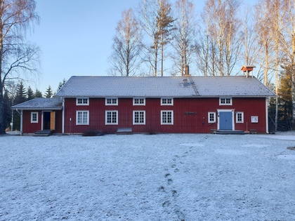 Röd trähus med vita fönster står på en snötäckt gräsmatta, omgiven av höga, kala träd.