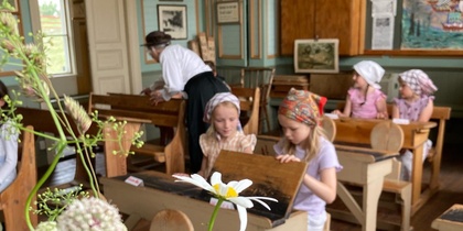 Barn i gammaldags kläder sitter vid bänkar i ett historiskt klassrum med blommor i förgrunden.