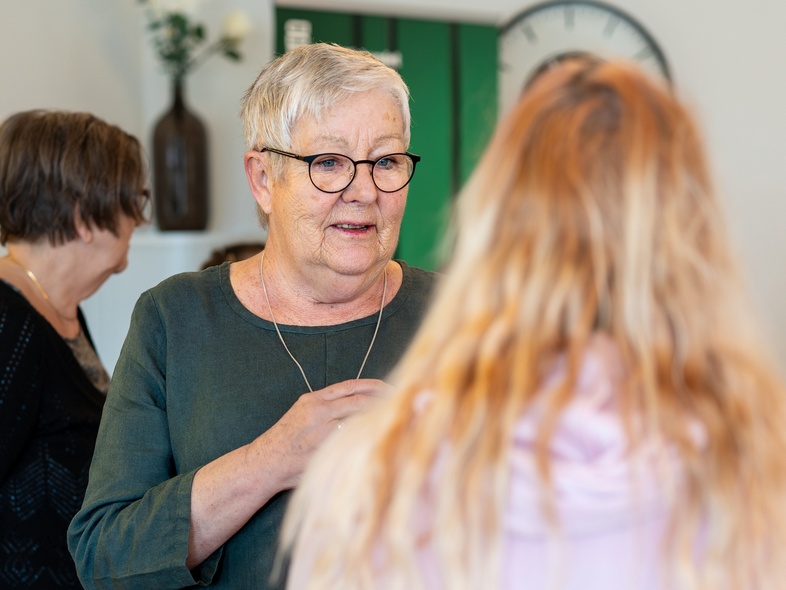 Äldre kvinna med glasögon samtalar med en yngre person i ett ljust rum med växter och klocka på väggen.