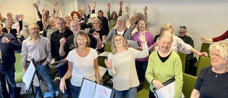 En grupp glada vuxna står i ett rum, vinkar och håller papper, troligen under en körövning.