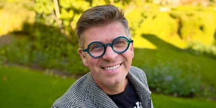 Person i rutigt kavaj och svart t-shirt står utomhus i en grönskande park.