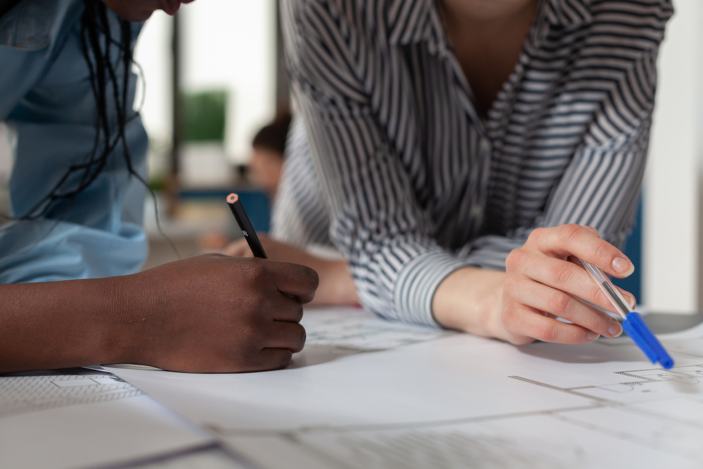 Två personer arbetar tillsammans vid ett bord och skriver på papper med penna och bläckpenna.