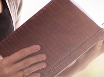 En person håller och läser en bok med brun pärm, endast överkroppen och händerna syns.