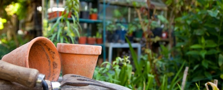 Trädgårdsredskap och lerkrukor på ett bord i en grönskande trädgård med växthus i bakgrunden.
